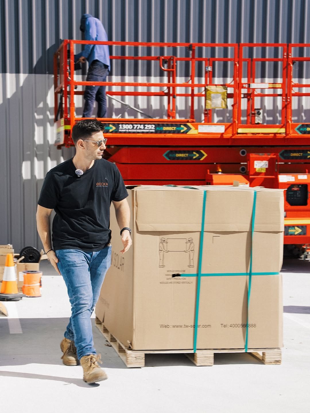 A Becka Electrical team member on a commercial site, walking past palletised solar modules with a scissor lift behind.