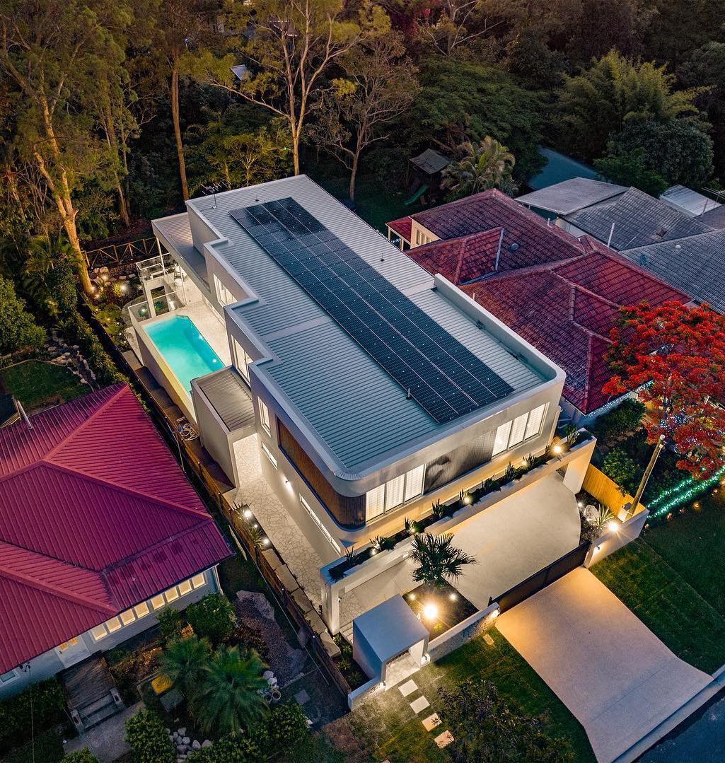 Aerial view at dusk of a Coorparoo home with a rooftop solar array, its pool lit and the driveway lights on.