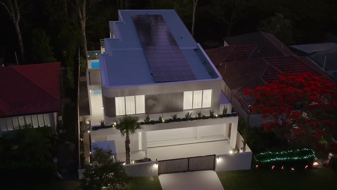Aerial view at dusk of a three-storey house with a rooftop solar array, lit pool and the surrounding street.