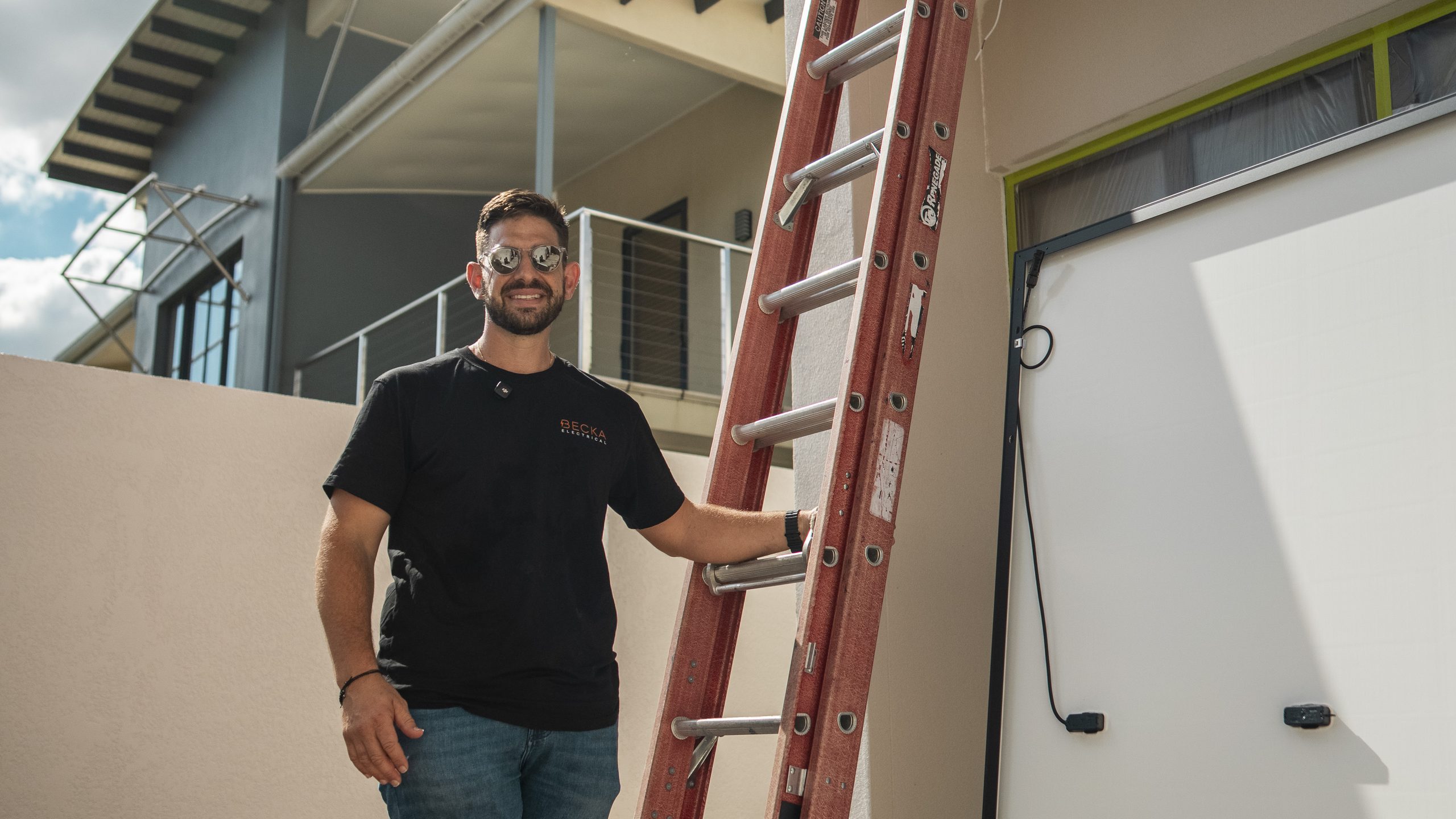 Becka Electrical electrician smiling at the camera, one hand on a ladder, beside a solar panel at a job site.