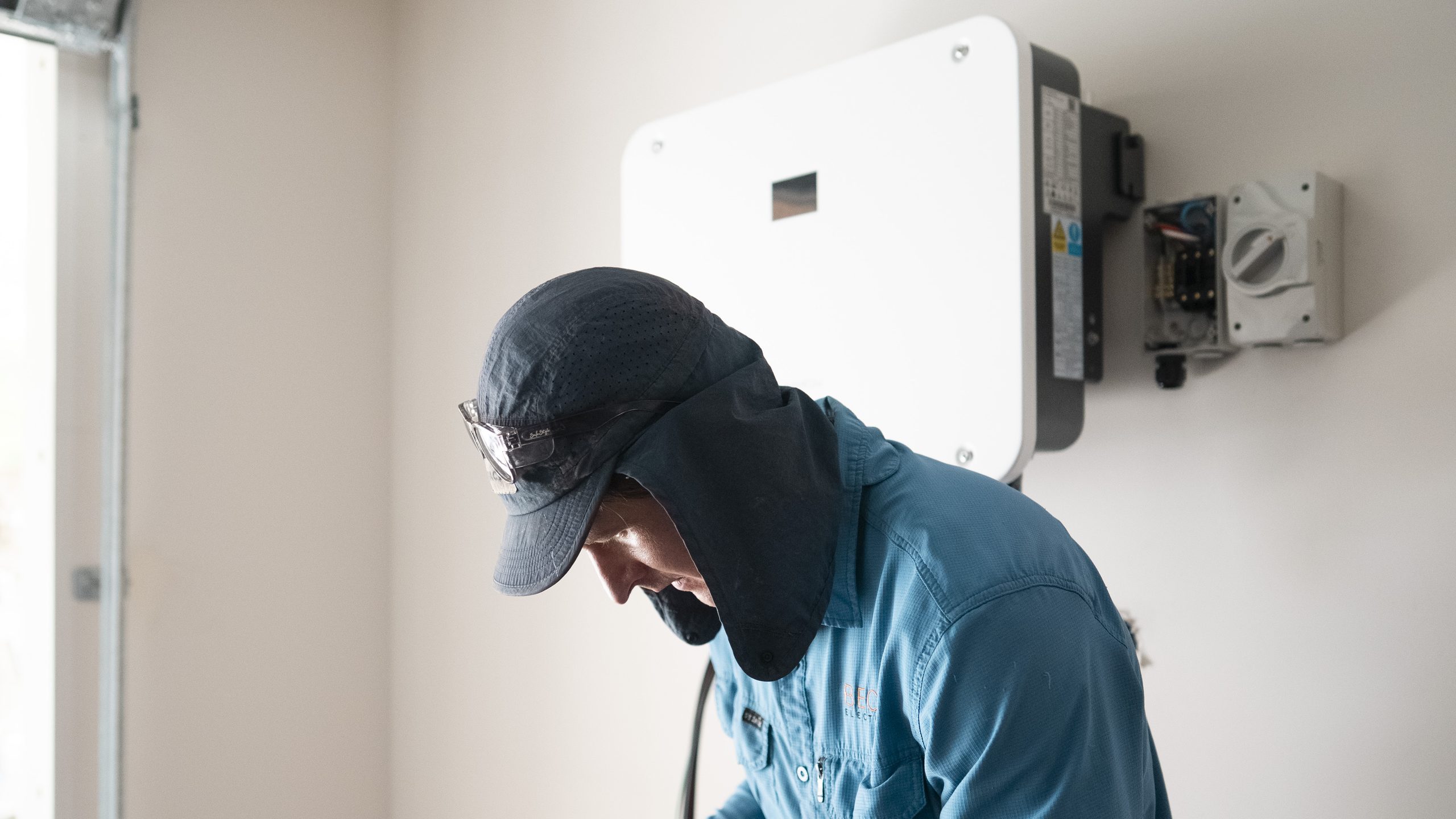 A Becka Electrical electrician leaning in to work at a wall-mounted solar inverter in a garage.