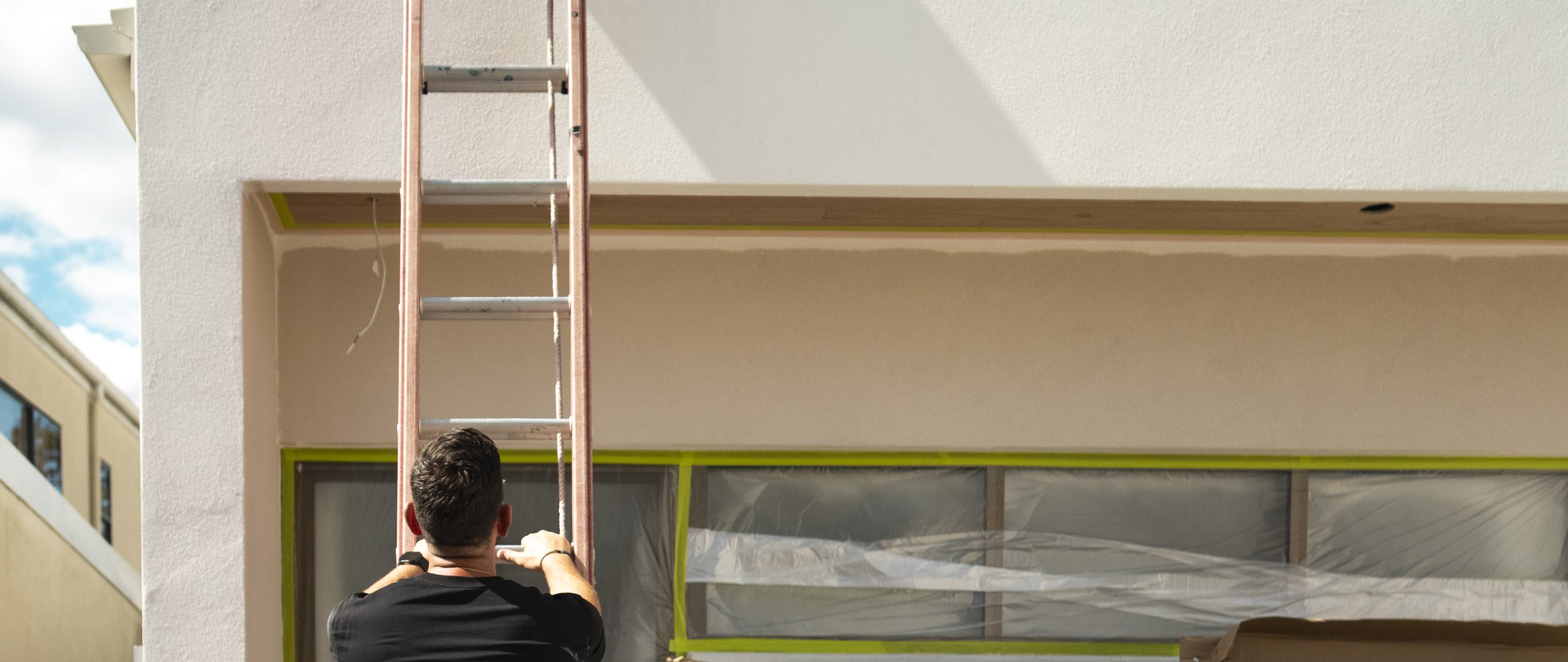 Becka electrician at the foot of an extension ladder against a rendered wall, with the windows taped and sheeted for work.