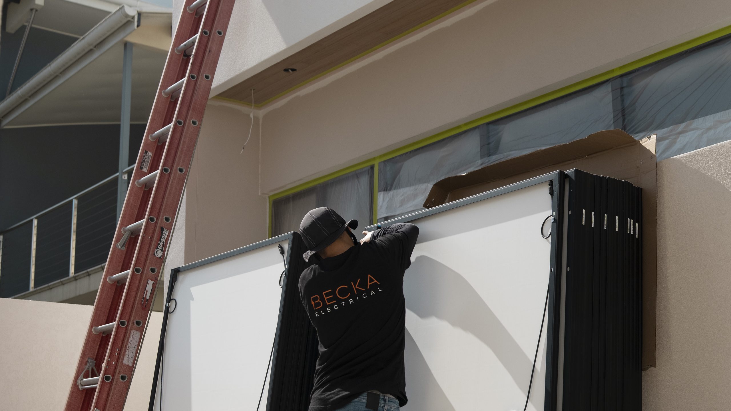 Becka electrician steadying a solar panel beside stacked panels at a job site.